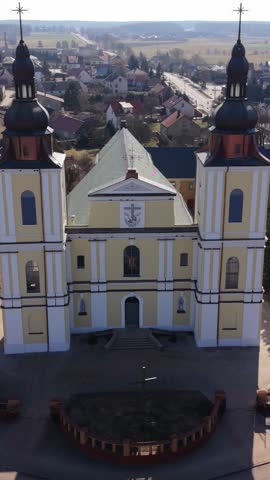 Vertical screen Close Aerial Of Church Front Facade Revealing Decorative Molding, Window Arches And Twin Bell Towers, Stone Steps And Semicircular Planting Bed Visible Under Warm Daylight