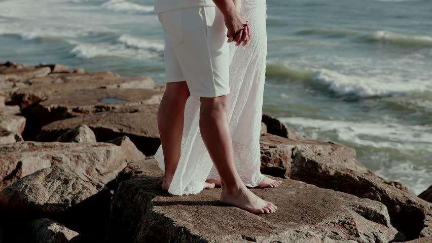 Man and woman in white clothing stand barefoot on coastal rocks facing ocean waves