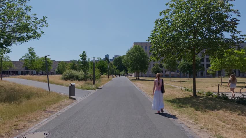 Woman walks along wide paved campus path framed by modern buildings, trees, dry grass, and distant skyline, creating calm urban university atmosphere with bright clear daylight and open space