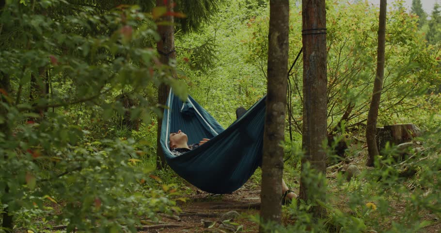 A man in sunglasses enjoys a peaceful nap in a blue hammock suspended between trees in a vibrant, ecofriendly forest setting.