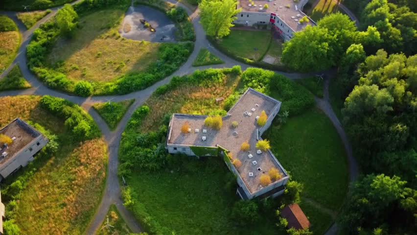 An aerial shot shows abandoned buildings surrounded by thick green vegetation and pathways. The buildings have flat roofs and trees growing nearby. This location appears to be abandoned for some time.
