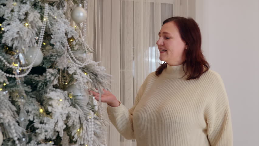 Woman wearing cream sweater smiles while looking at heavily frosted Christmas tree decorated with white ornaments, golden lights in modern home setting.