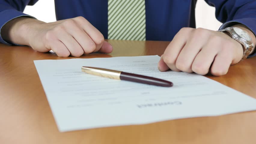 A closeup of a man in a blue shirt and striped tie signing a document on a wooden table