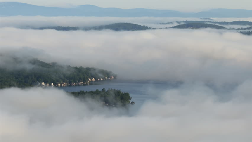 Telephoto aerial view of morning fog covering much of Lake Sunapee as it opens over Hedgehog Cove, revealing waterfront homes and a wooded shoreline beneath layered distant hills looking westward