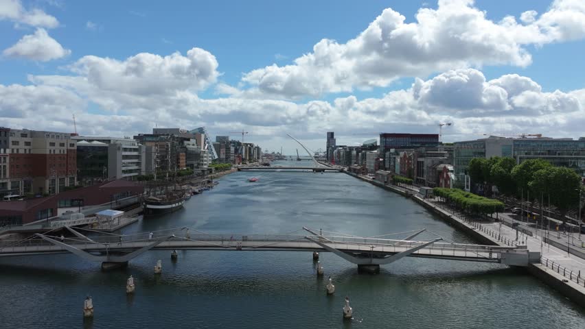 Camera glides along the Liffey past modern docklands, twin pedestrian bridge and Samuel Beckett Bridge on a breezy summer day.