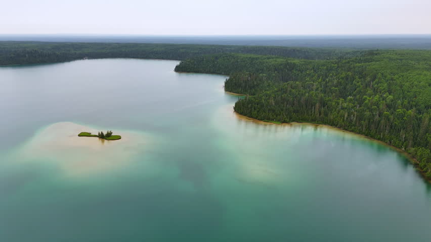 Aerial view of a grass-covered sandy island surrounded by still turquoise water at Big Skunk Lake in Northern Ontario, with dense boreal forest extending to the horizon.