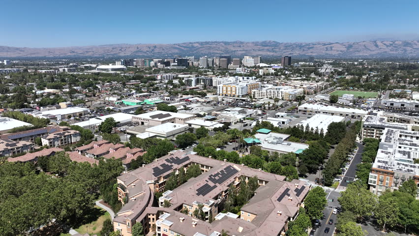 Wide aerial drone view of downtown San Jose, California, featuring skyline buildings, surrounding residential neighborhoods, and the Diablo Range mountains in the background on a clear sunny day.