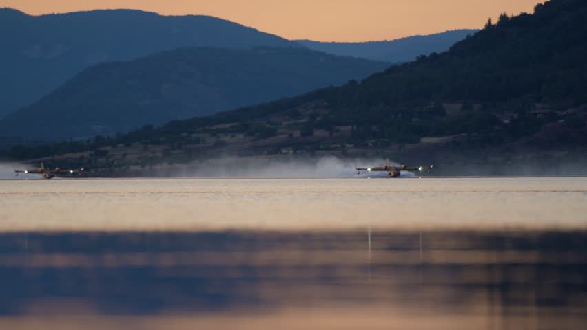 Firefighting planes skim a calm lake, scooping water at golden dusk.