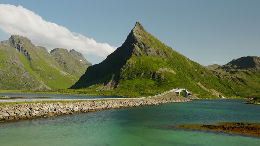 A low causeway and arch bridge cross turquoise water toward a sharp rocky peak under clear summer skies in Norway Lofoten.
