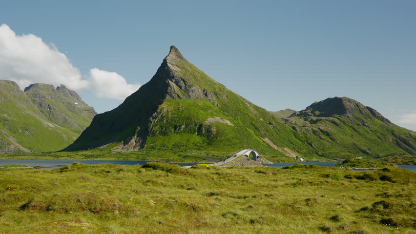 Jagged peak, arched bridge, and blue inlet sit beyond open grassy tundra under clear summer skies.