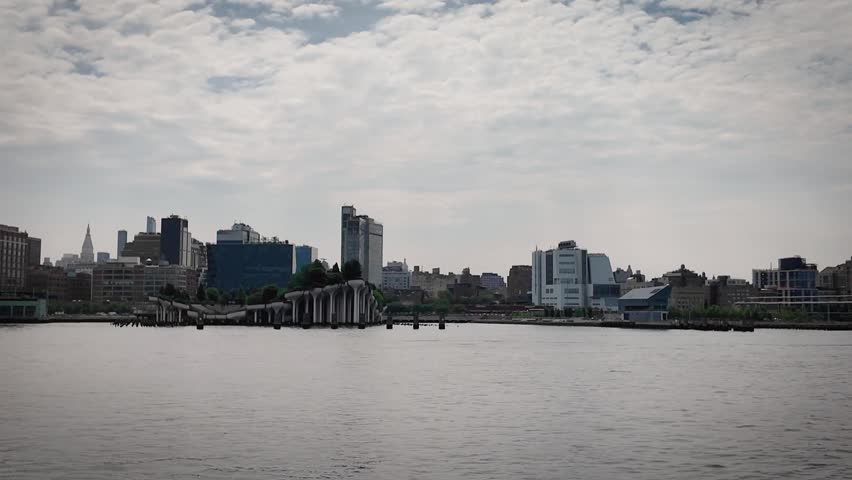 Boat-level view of Little Island on the Hudson River featuring its distinctive elevated concrete columns, waterfront buildings and the Manhattan skyline beneath a dramatic cloudy sky.