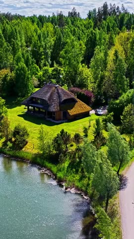 Aerial view of a traditional house with a thatched roof nestled in a lush forest by a lake in Amatciems, Cesu novads, Latvia.