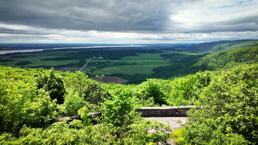 Wide panoramic landscape from Champlain Lookout in Gatineau Park overlooking the Ottawa Valley with forests, farmland and distant waterways beneath dramatic storm clouds and changing sunlight.