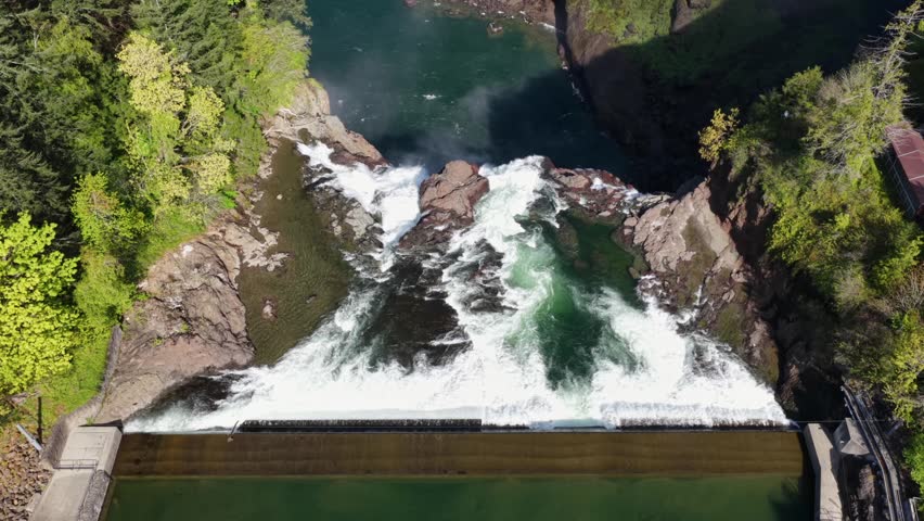Snoqualmie Falls cascades powerfully over rugged rocky cliff, churning white water plunges into deep turquoise pool surrounded by dense green Washington forest canopy. Aerial drone top down flyover