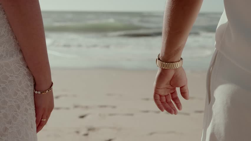 Close up of clasped hands with bracelet and watch as couple walks along shoreline sand