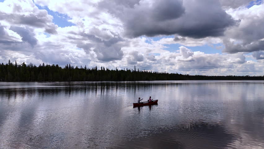 Drone follows a couple paddling a canoe across a calm wilderness lake beneath bright clouds, capturing a peaceful outdoor adventure in Northern Ontario.