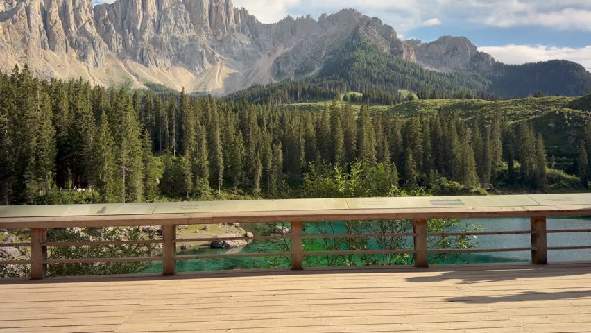 A wooden boardwalk with an informational sign along a nature trail overlooks Lake Carezza, with the Dolomite mountains rising beyond the forest, South Tyrol, Italy.