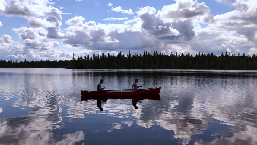 A couple begins a canoe trip across a calm lake reflecting clouds beneath a bright sky, surrounded by peaceful forest in the Northern Ontario wilderness.