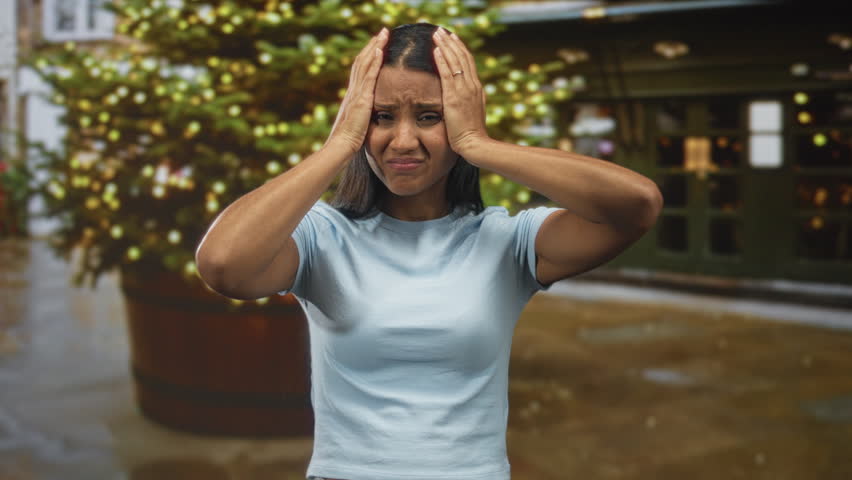 Woman holding head with both hands, eyes closed and wincing near a lit holiday tree and shopfront on a wet street; stress fatigue.