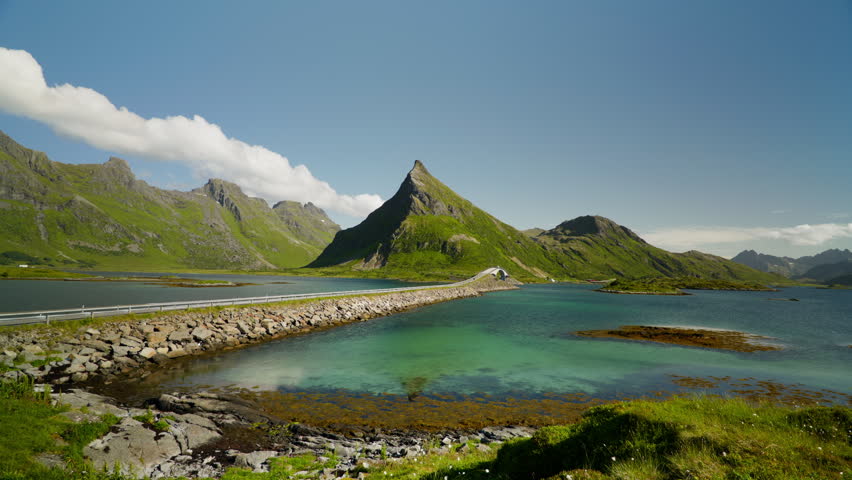 Rocky causeway crosses a turquoise bay toward a sharp grass covered peak under clear blue summer skies in northern Norway.