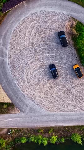 Aerial view of cars parked on a gravel lot surrounded by lush green trees and a calm lake in Amatciems, Cesu novads, Latvia.