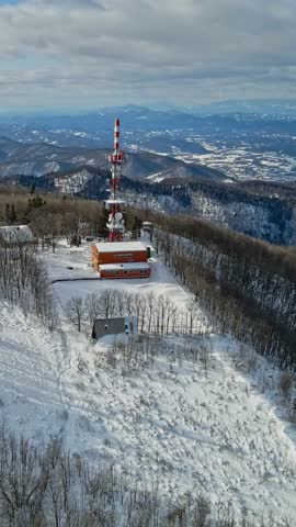 Vertical video aerial footage of a red and white broadcast tower on a snowy mountain, surrounded by frosty trees and panoramic winter landscape.