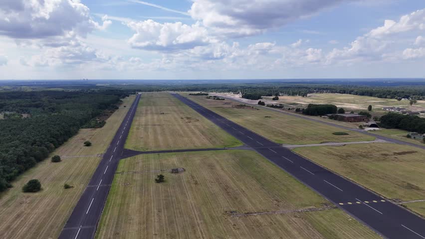 An Aerial shot of a long, dark asphalt runway cutting through a wide grass field under a cloudy sky