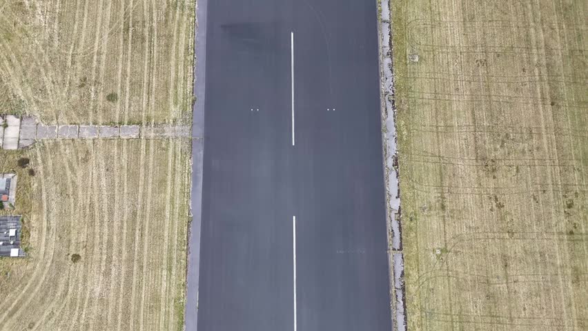 An Aerial shot of a long, dark asphalt runway cutting through a wide grass field under a cloudy sky