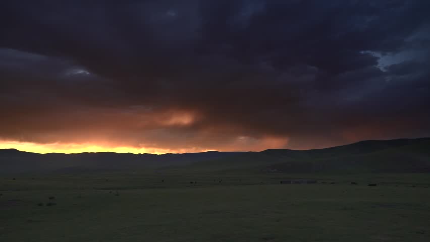 Mongolia sunset glows above grassland hills beneath dramatic storm clouds, evening color. Twilight spreads across rolling plains with moody atmosphere, distant mountains. Warm sunset colors appear as low-angle sunlight passes through a longer section of the atmosphere and scatters cooler wavelengths. Dense storm clouds create tonal contrast above the illuminated horizon and emphasize the changing weather of a continental grassland. The layered hills, open sky, and fading daylight provide atmospheric imagery for nature, climate, solitude, travel, and environmental backgrounds.