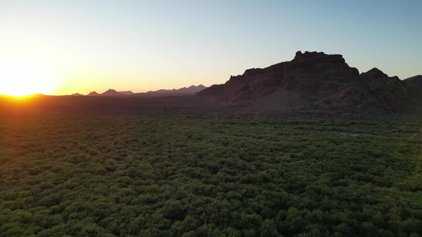 A drone view of desert mountain peak looms over a large field of green brush under twilight sky in the Sonoran Desert, Mesa, Arizona