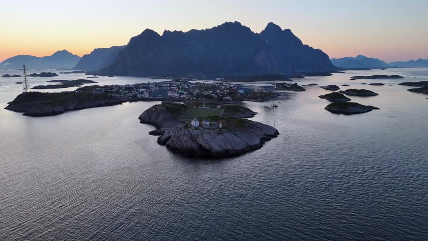 An aerial view of the Norwegian fishing village of Henningvaer illuminated by the soft glow of the midnight sun, with the towering mount Festvagtind