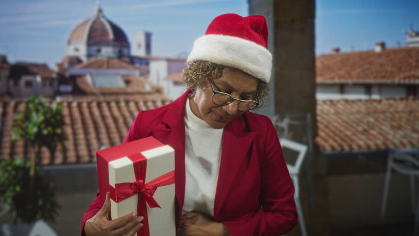 Woman holding wrapped gift box with hand on chest in building rooftop overlooking tiled roofs and a distant dome; nostalgia.
