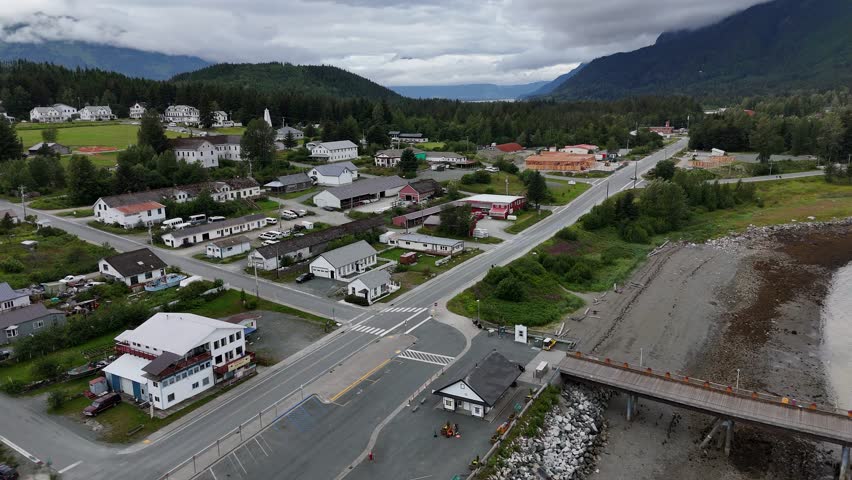 An aerial view shows a small coastal town with a grid of streets and houses and lush mountains in the distance under a cloudy sky in Haines, Alaska