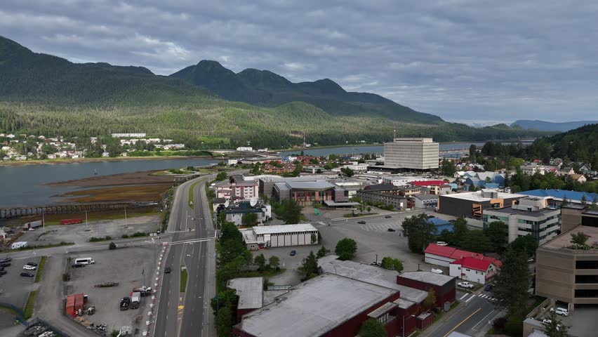 A drone view above Juneau, Alaska, showcasing the port with water, highway, buildings and green mountains under cloudy sky