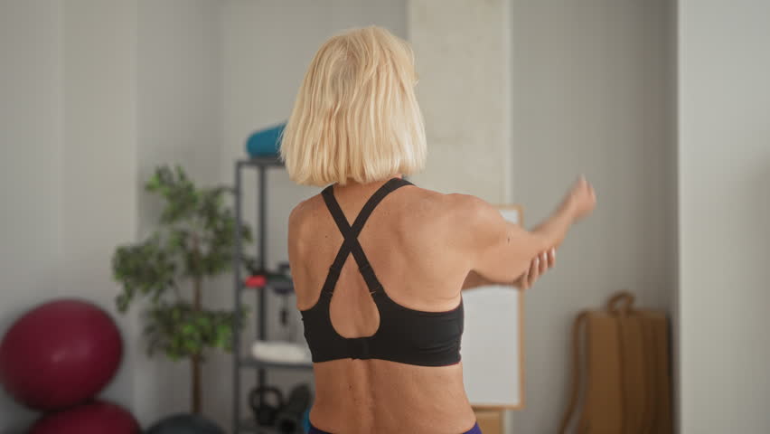 Middle age blonde woman reaching right hand behind back to touch left shoulder and stretch triceps while training in a yoga studio; calm wellbeing.