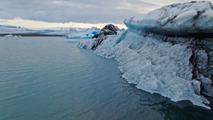 Cinematic aerial flight above floating blue icebergs in Jökulsárlón Glacier Lagoon, Iceland, revealing the breathtaking beauty of Vatnajökull glacier and the Arctic landscape at sunset.