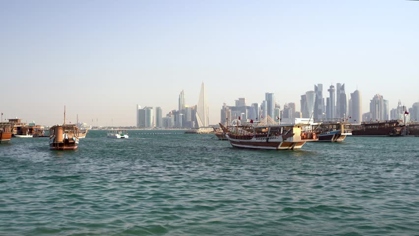 A scenic wide shot across the blue waters of Doha Bay, showcasing traditional wooden dhow boats resting in the harbor, beautifully contrasted against the towering, modern skyscrapers of the Doha city skyline in the distance on a bright, clear day.