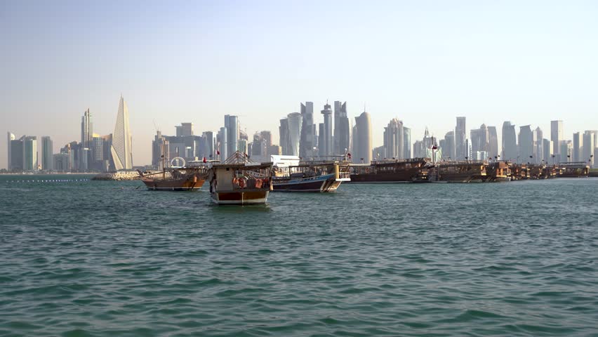 A scenic panoramic view of Doha Bay, featuring a collection of traditional wooden dhow boats moored in the foreground contrasted against the modern, towering skyscraper skyline of Doha, Qatar, on a bright day.