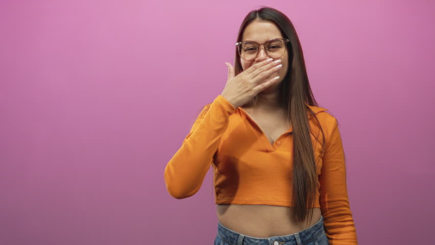 Woman covering mouth with hand in studio, wearing orange crop top and jeans, glasses and long brunette hair against pink backdrop; amused candid moment.