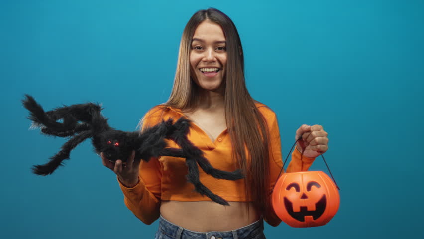 Woman holding orange pumpkin bucket and large fuzzy spider with outstretched hands against studio backdrop; festive fun.