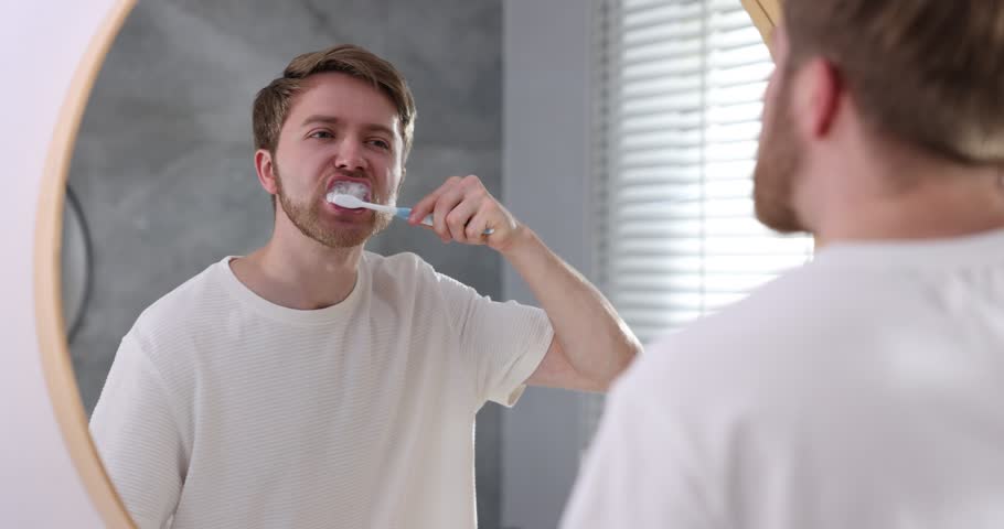Man suffering from pain while brushing teeth near mirror in bathroom. Oral hygiene
