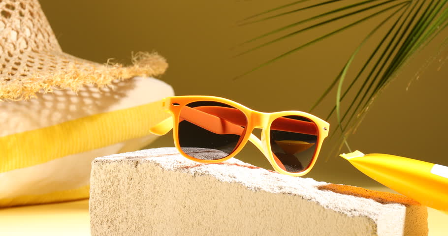 Stylish sunglasses, sunscreen, blanket, hat and decor on pale yellow background, closeup. Changing direction of light