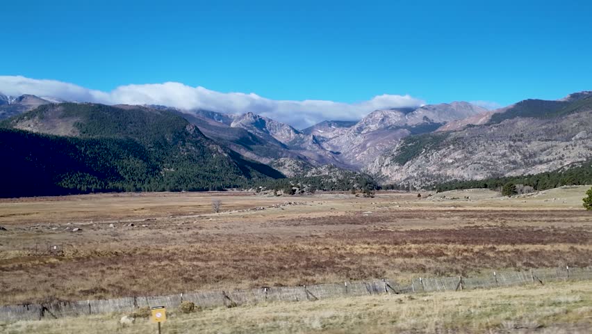 A scenic driving shot across open meadows toward the towering Rocky Mountains near Estes Park, Colorado beneath clear blue skies and rolling clouds.