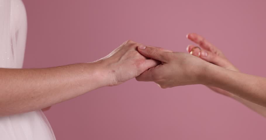 Lesbian wedding. Women putting rings on each others fingers against pink background, closeup