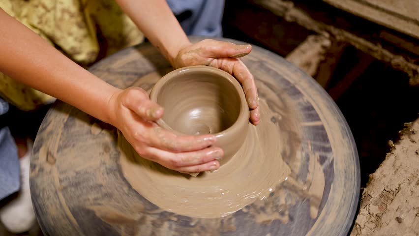 Close-up overhead view of potter molding wet clay into ceramic bowl on spinning wheel.