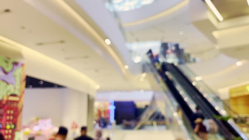A softly blurred shopping mall interior features an escalator, bright ceiling lights, retail activity, and moving customers in a modern space.