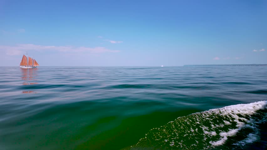 The wake of a scenic boat tour drifts across the crystal-clear waters of Green Bay before revealing a red sailboat gliding beneath the summer sky near Sister Bay, Wisconsin.