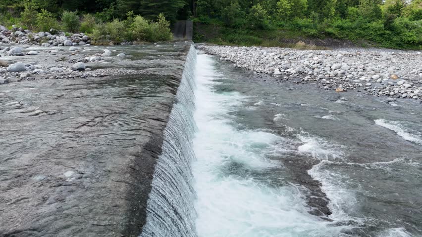 Clear water flows over a small dam. Rocks line both sides of the stream. Green trees surround the riverbank. Nature