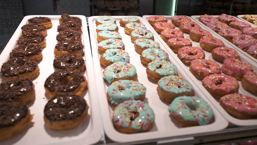 Rows of iced donuts with sprinkles line white trays in a bakery display inside a food store.