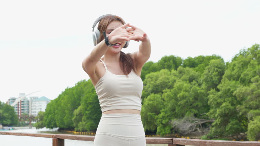 Young woman stretching before run for healthy lifestyle and sport smiles while wearing wireless headphones and smartwatch during outdoor workout session near green forest background.